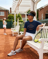 Woman sitting on a bench with a tennis racket on a tennis court, another player in the background.