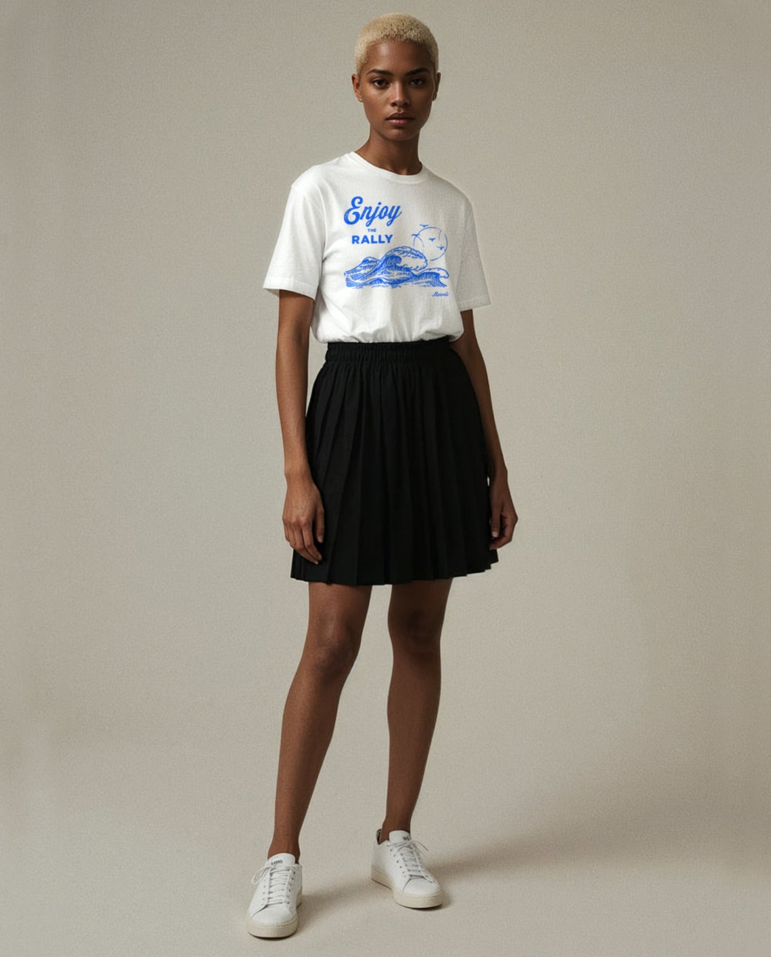 Person wearing a white rally t-shirt with tennis and surf graphics and text, black skirt, and white sneakers