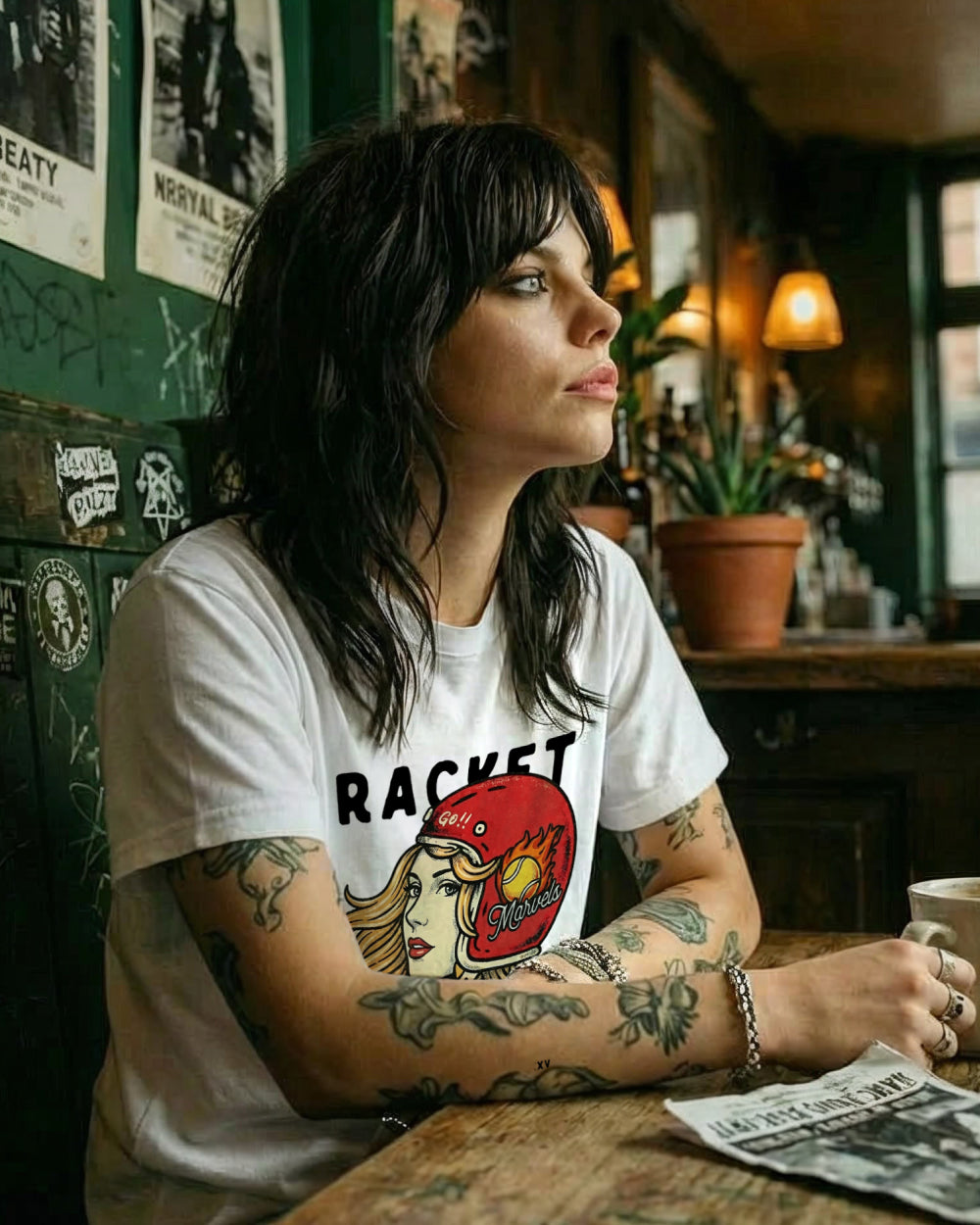 Woman with tattoos wearing a white t-shirt with a graphic design, sitting at a table in a bar.