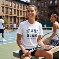 Woman on a tennis court wearing a white t-shirt with 'Grand Slam' text, holding a tennis racket.
