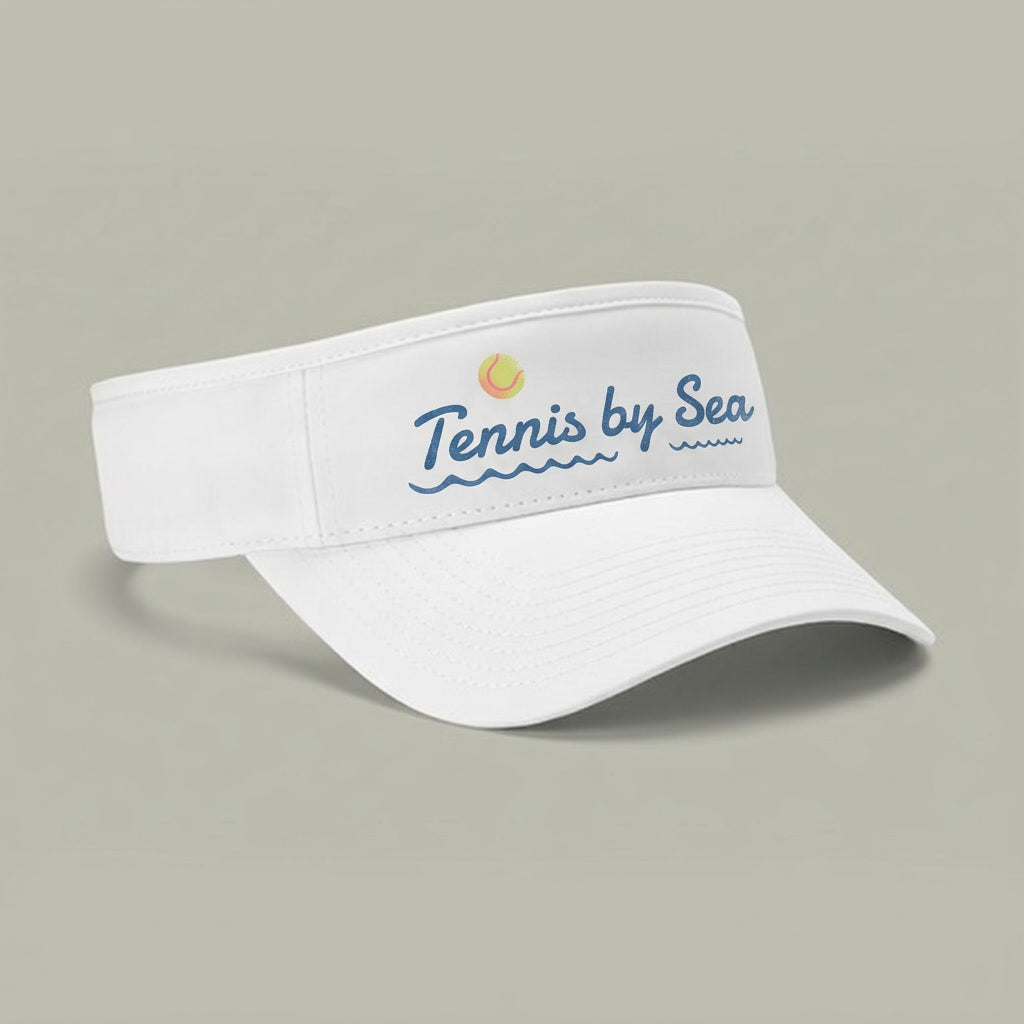 A white visor hat with 'Tennis by Sea' printed in blue script on the front, accompanied by a small yellow tennis ball graphic, set against a solid gray background.