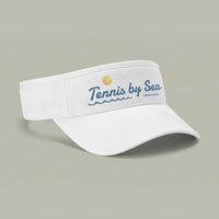 A white visor hat with 'Tennis by Sea' printed in blue script on the front, accompanied by a small yellow tennis ball graphic, set against a solid gray background.