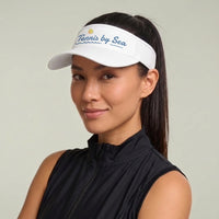 A woman wearing a white visor with 'Tennis by Sea' embroidered in blue and yellow, paired with a black sleeveless athletic top, against a light gray background.